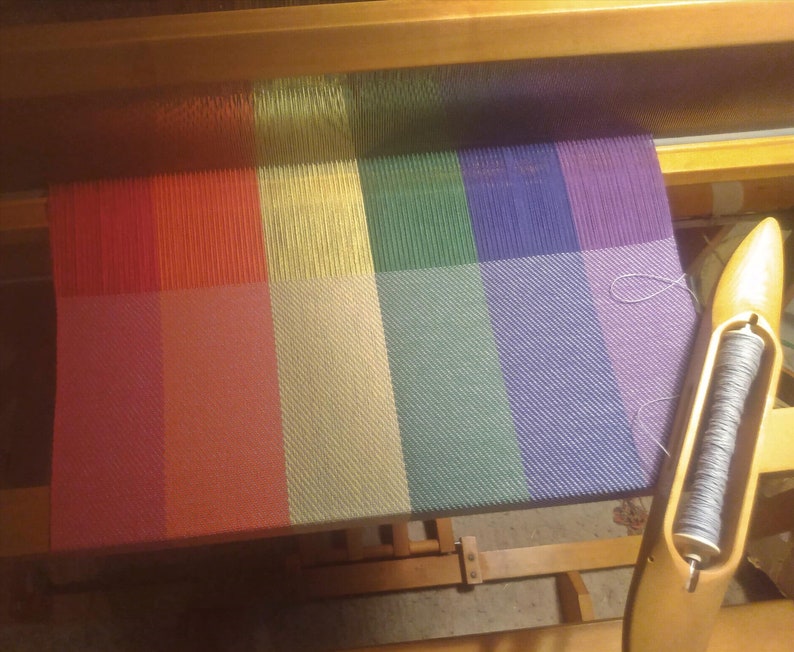 The fabric is shown being woven on a four-harness Harrisville loom. The shuttle holding the light silver weft yarn sits at an angle on the lower right corner of the fabric.