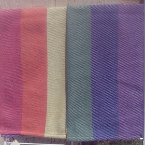 Two folded handwoven rainbow pride hand towels hang side by side, one showing red, orange, and yellow stripes, the other green, blue, and purple stripes. Each stripe is just shy of 3 inches wide.