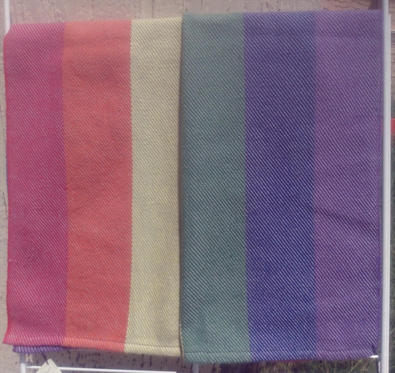 Two folded handwoven rainbow pride hand towels hang side by side, one showing red, orange, and yellow stripes, the other green, blue, and purple stripes. Each stripe is just shy of 3 inches wide.