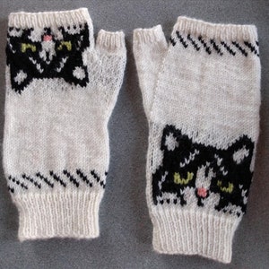 Könnte beinhalten: Ein Paar weiße gestrickte Fingerlose Handschuhe mit schwarzen und grünen Katzenköpfen. Die Handschuhe haben ein schwarz-weiß gestreiftes Muster am Handgelenk.