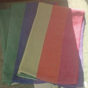 Two folded handwoven rainbow pride hand towels are shown, one lying partially atop the other. Each stripe is just shy of 3 inches wide.