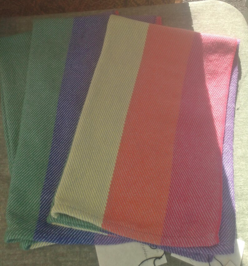 Two folded handwoven rainbow pride hand towels are shown, one lying partially atop the other. Each stripe is just shy of 3 inches wide.