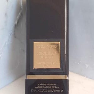 Può includere: Un flacone di profumo rettangolare nero con un'etichetta dorata. L'etichetta recita "TOM FORD JONQUILLE DE NUIT EAU DE PARFUM" e "50 ml". Il flacone è su una superficie bianca e grigia.