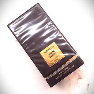 Può includere: Scatola rettangolare marrone scuro di Tom Ford White Suede Eau de Parfum, avvolta in plastica trasparente. Un'etichetta dorata mostra il nome del prodotto e il volume: 250 ml.