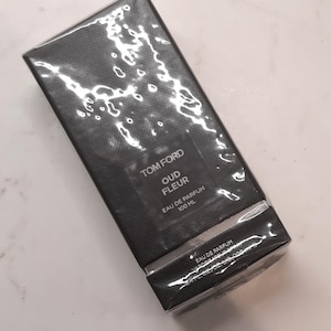 Può includere: Una scatola nera con un'etichetta argentata che recita "TOM FORD OUD FLEUR EAU DE PARFUM 100 ML". La scatola è avvolta in plastica trasparente.