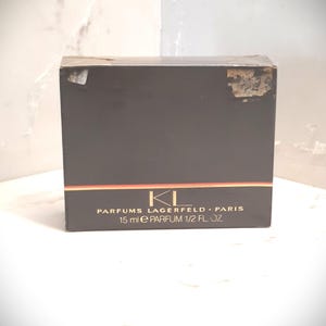 Op de afbeelding: Een zwarte doos met gouden letters die "Parfums Lagerfeld - Paris" en "15 ml e Parfum 1/2 Fl. Oz." zeggen.