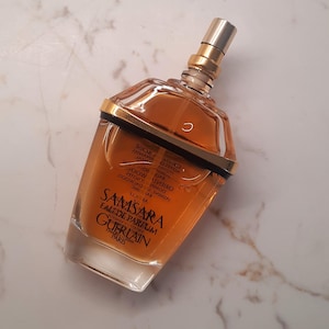 Puede incluir: Una botella de vidrio transparente de Samsara Eau de Parfum de Guerlain. El frasco de perfume tiene una tapa dorada y una banda dorada alrededor de la parte superior. El líquido del interior es de color ámbar. La botella está sobre una superficie de mármol blanco y gris.