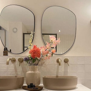 May include: Two oval-shaped mirrors with black frames hang above a white bathroom vanity with two brown ceramic sinks. A vase with pink and red flowers sits between the sinks. The vanity has a white drawer and a white countertop.