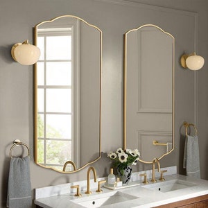 May include: Two arched mirrors with gold frames hang above a white double vanity with two sinks and gold taps. A white towel hangs on a gold ring on the left side of the vanity. A bouquet of white flowers sits on the counter between the sinks.