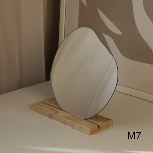 May include: A freeform, organic-shaped mirror with a silver backing sits on a wooden stand. The stand is made of light brown wood with a natural grain. The mirror is on a white surface. M7