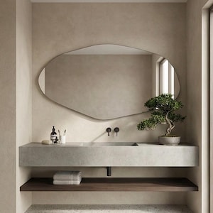 May include: A modern bathroom scene featuring a concrete vanity with a built-in sink and a dark wood shelf below. An irregularly shaped mirror hangs above the vanity. A small bonsai tree sits on the right, adding a touch of nature.