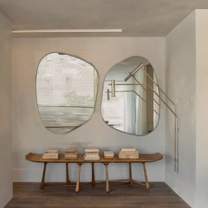 May include: An interior shot featuring two irregularly shaped mirrors mounted on a light gray wall. Below the mirrors is a wooden bench with several small, square wooden boxes. The floor is a light brown wood.
