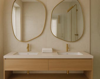 May include: A modern bathroom vanity with two sinks, gold fixtures, and two irregularly shaped mirrors. The vanity is made of light wood with a white countertop and a lower shelf. A white towel is placed between the sinks.