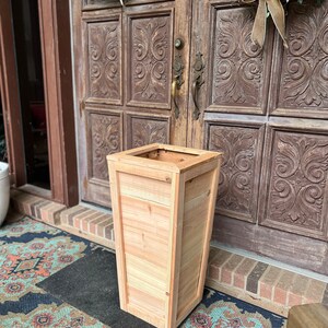 May include: A natural wood planter with a rectangular shape and a slightly angled top. The planter is sitting on a patterned rug in front of a wooden door with decorative carvings.