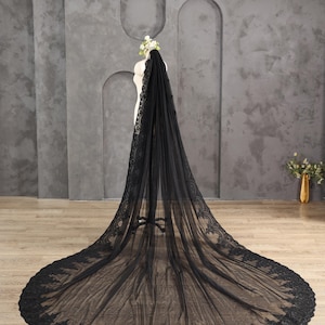 May include: A long, black bridal veil with intricate lace detailing along the edges. The veil is displayed on a mannequin, showcasing its flowing design and delicate texture. The veil's train extends across the floor.