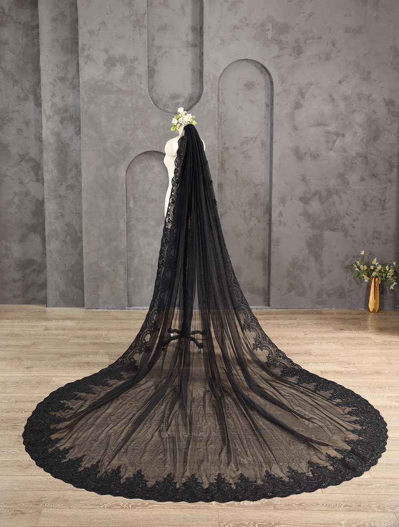 May include: A long, black bridal veil with intricate lace detailing along the edges. The veil is displayed on a mannequin, showcasing its flowing design and delicate texture. The veil's train extends across the floor.