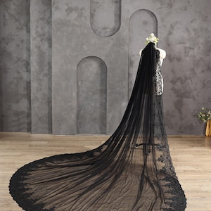 May include: A black wedding veil with a long train, displayed on a mannequin. The veil is made of sheer fabric and features intricate lace detailing along the edges. A floral headpiece adorns the mannequin's head, complementing the veil's design.