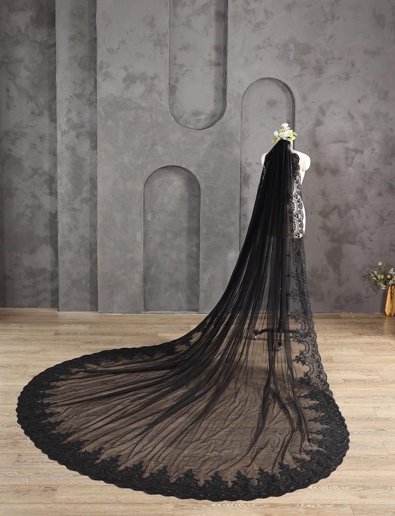 May include: A black wedding veil with a long train, displayed on a mannequin. The veil is made of sheer fabric and features intricate lace detailing along the edges. A floral headpiece adorns the mannequin's head, complementing the veil's design.