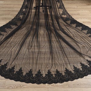 May include: A long, black veil with intricate, ornate lace detailing along the edges. The veil is made of a sheer fabric, creating a delicate and elegant appearance. The lace design features a repeating floral pattern.