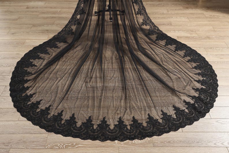 May include: A long, black veil with intricate, ornate lace detailing along the edges. The veil is made of a sheer fabric, creating a delicate and elegant appearance. The lace design features a repeating floral pattern.