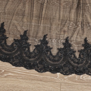 May include: Black lace trim with a scalloped edge and ornate floral and scroll designs. The lace is set against a wooden background. This trim is suitable for various sewing and craft projects.