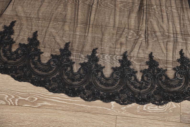 May include: Black lace trim with a scalloped edge and ornate floral and scroll designs. The lace is set against a wooden background. This trim is suitable for various sewing and craft projects.