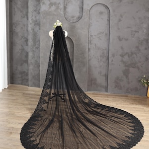 May include: A long, black bridal veil with intricate lace detailing along the edges. The veil is displayed on a mannequin, showcasing its flowing design and delicate floral crown. The veil's train extends across the floor.