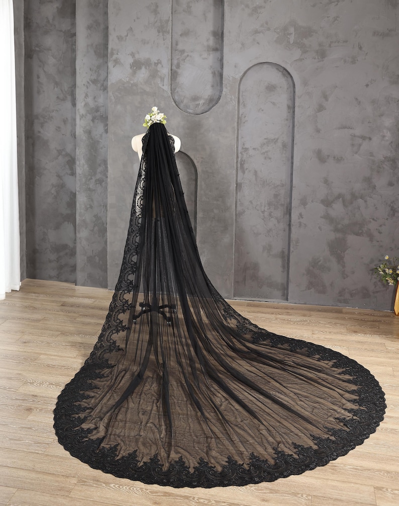 May include: A long, black bridal veil with intricate lace detailing along the edges. The veil is displayed on a mannequin, showcasing its flowing design and delicate floral crown. The veil's train extends across the floor.
