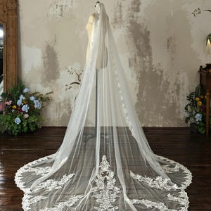 May include: A long, floor-length ivory wedding veil draped over a mannequin. The sheer tulle fabric features intricate white floral lace embroidery along the edges and a large, detailed lace applique panel at the base of the train.