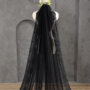 May include: A black bridal veil with intricate lace detailing along the edges. The veil is draped over a mannequin, showcasing its length and design. A small floral arrangement sits atop the mannequin's head, complementing the veil's elegance.