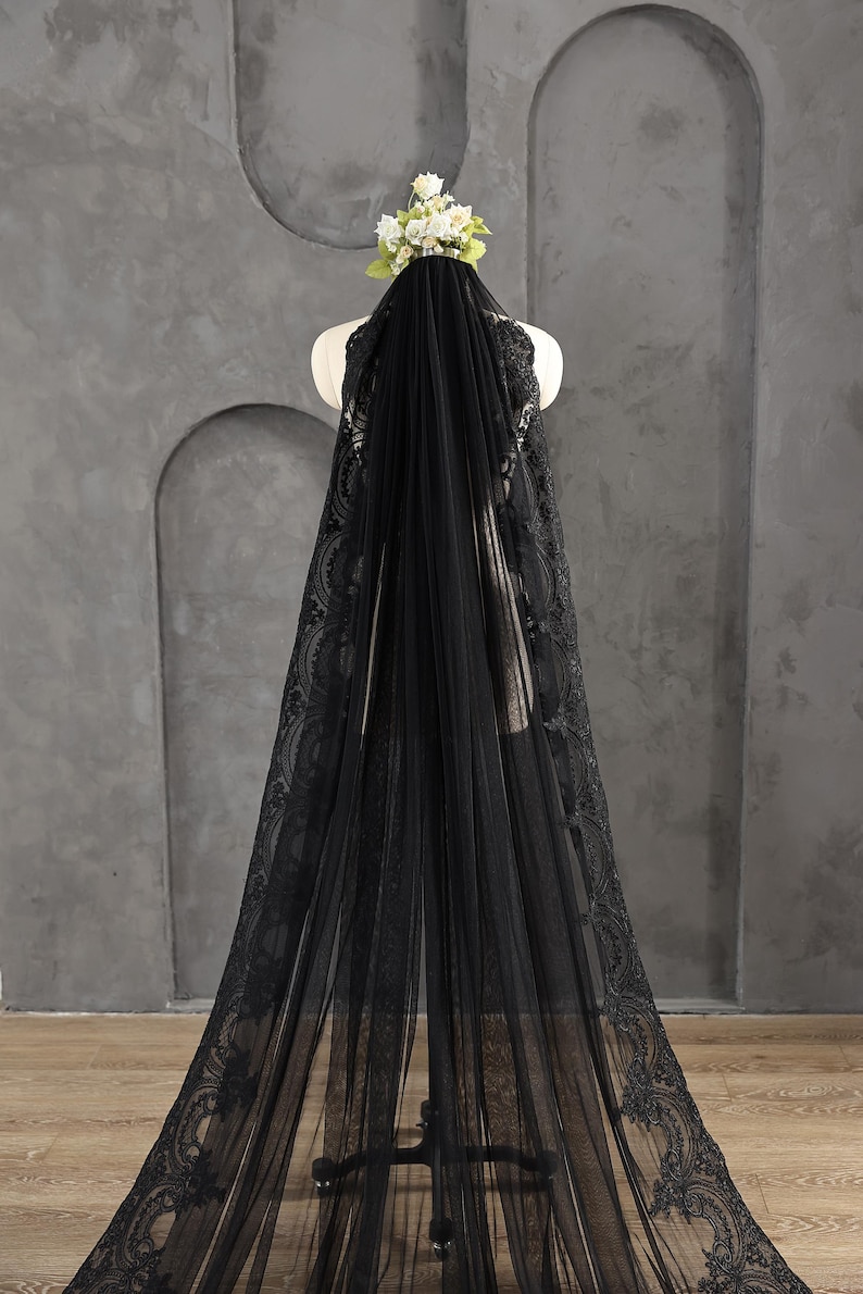 May include: A black bridal veil with intricate lace detailing along the edges. The veil is draped over a mannequin, showcasing its length and design. A small floral arrangement sits atop the mannequin's head, complementing the veil's elegance.