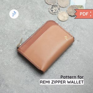 Könnte beinhalten: Eine braune Ledergeldbörse mit Reißverschluss und einem kontrastierenden, hellbraunen Lederpanel. Die Geldbörse hat ein kleines Logo auf der Vorderseite und wird mit einem Reißverschluss geschlossen.