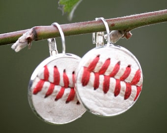Puede incluir: Un par de pendientes de béisbol plateados con costuras rojas. Los pendientes están colgados de una rama.