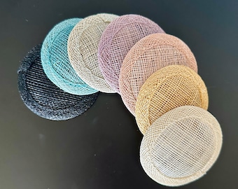 May include: A collection of seven circular fascinator bases arranged in a fan shape. The hats feature a woven mesh texture and come in black, teal, light gray, mauve, dusty rose, gold, and cream colors, each with a raised, rounded edge.