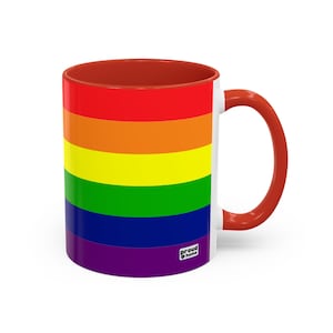 May include: A white ceramic mug with a red handle and a rainbow flag design. The mug has a red interior and a small "proud home" logo on the bottom.