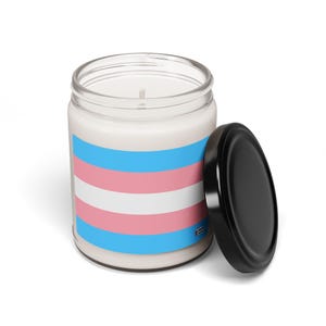 Op de afbeelding: Een glazen kaars pot met een wit deksel. De kaars is versierd met de transgender pride-vlag, die lichtblauwe, roze en witte horizontale strepen heeft.