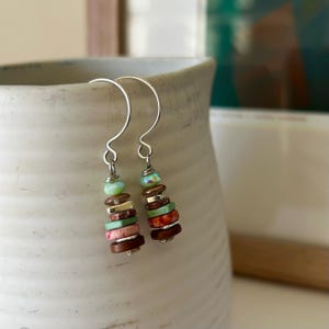 Puede incluir: Un par de pendientes hechos a mano con ganchos plateados. Cada pendiente presenta una pila de cuentas coloridas, incluyendo verde, rosa, marrón y rojo, creando un efecto en capas. Las cuentas tienen una mezcla de texturas y acabados.