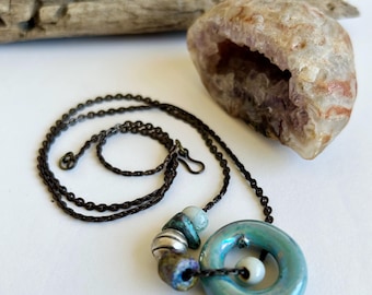 May include: A turquoise blue ceramic pendant necklace with a swirl design. The pendant is accented with silver and blue beads. The necklace is on a dark brown chain.