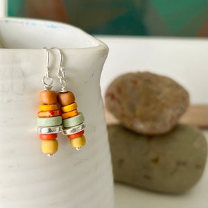 Peut inclure: Une paire de boucles d'oreilles pendantes avec une pile de perles colorées dans des tons d'orange, de jaune et de vert. Les perles sont séparées par des entretoises argentées. Les boucles d'oreilles sont faites avec des crochets argentés.