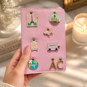 Könnte beinhalten: Rosa Reisepasshülle mit Reise-Pins, darunter ein Miniatur-Eiffelturm, ein Schloss und ein Doppeldeckerbus. Die Pins sind mit goldfarbenen Nieten befestigt. Der Text "Explore" ist auf einem der Pins zu sehen.