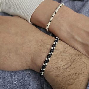 Könnte beinhalten: Zwei passende Armbänder. Eines ist schwarz und weiß, das andere weiß und schwarz. Die Armbänder sind aus geflochtenem Kordel gefertigt. Die Armbänder werden an den Handgelenken getragen.
