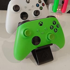 Puede incluir: Dos mandos de Xbox, uno blanco y otro verde brillante, están apilados uno encima del otro. El mando verde está en primer plano, con un soporte negro adjunto. Otros accesorios de juego son visibles en el fondo.
