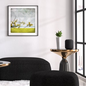 May include: A black and gold side table with a round top and a single leg. A black speaker and a potted plant are on the table. A black couch and ottoman are in the foreground. A framed print of a yellow flower with a blue and white background is on the wall.