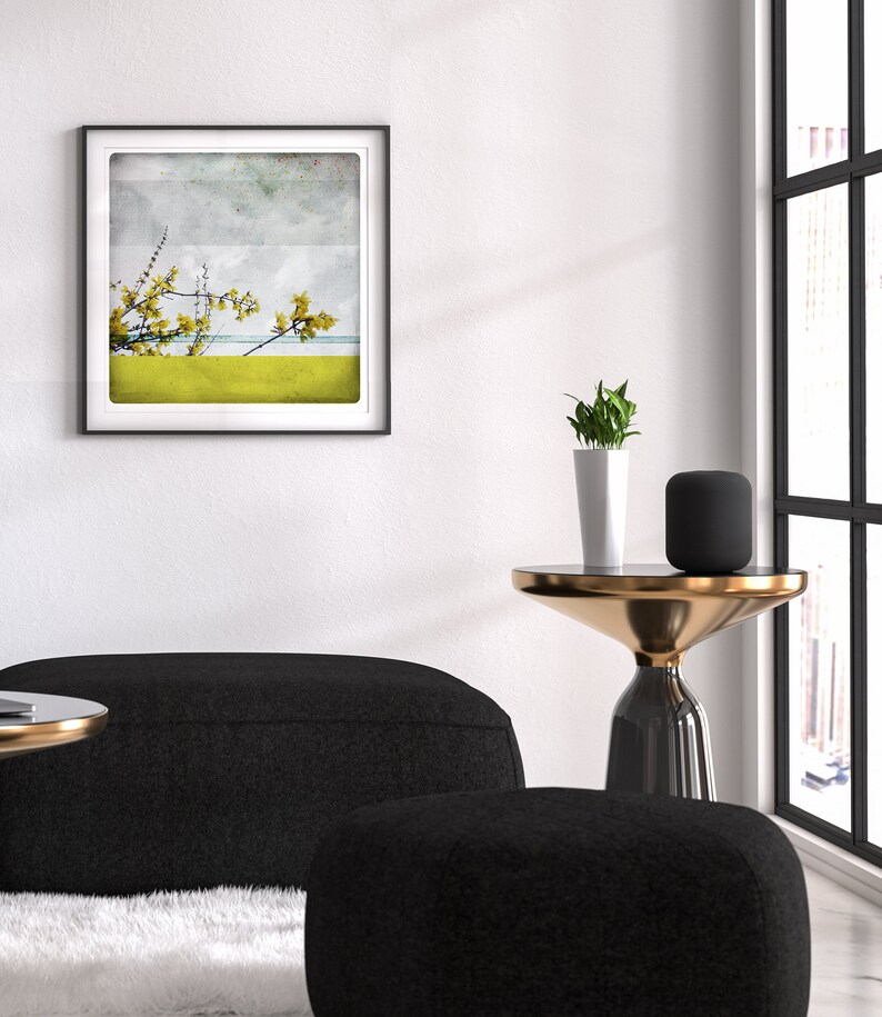 May include: A black and gold side table with a round top and a single leg. A black speaker and a potted plant are on the table. A black couch and ottoman are in the foreground. A framed print of a yellow flower with a blue and white background is on the wall.