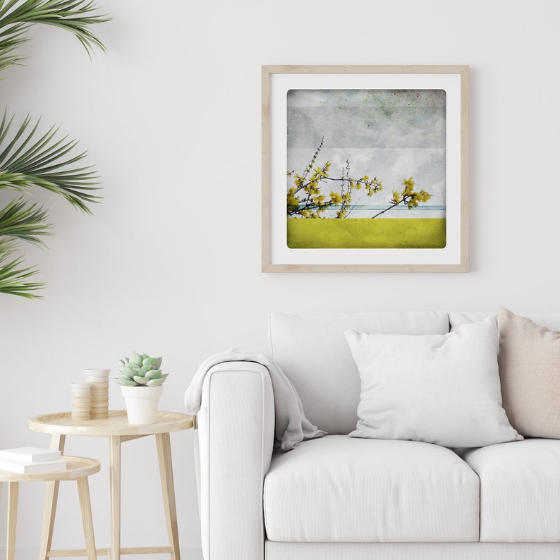 May include: A framed print of a watercolor painting depicting a yellow flowering branch against a blue and gray sky. The painting is framed in a light wood frame and is hanging on a white wall.