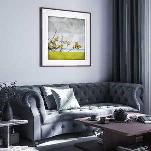 May include: A dark gray tufted sofa with a light blue throw pillow in a living room setting. A framed print of a yellow flower branch against a blue and gray background hangs on the wall. A wooden coffee table with a bowl of fruit and a magazine sits in front of the sofa.
