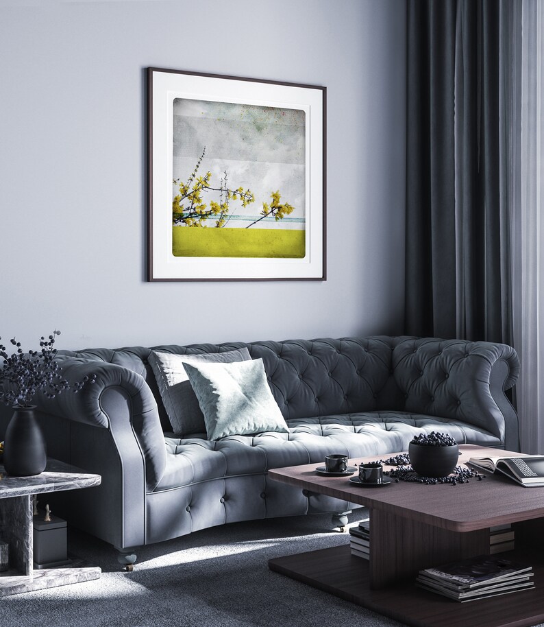 May include: A dark gray tufted sofa with a light blue throw pillow in a living room setting. A framed print of a yellow flower branch against a blue and gray background hangs on the wall. A wooden coffee table with a bowl of fruit and a magazine sits in front of the sofa.