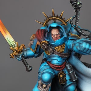 Könnte beinhalten: Detaillierte Miniaturfigur eines Kriegers in blauer Rüstung, der ein farbwechselndes Schwert hält. Die Figur hat einen strengen Ausdruck und ist mit goldenen Akzenten und einem fließenden Umhang verziert. Ein schädelbesetztes Gerät ist angebracht.