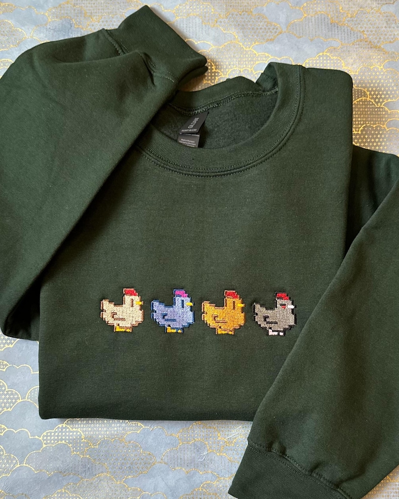 K&ouml;nnte beinhalten: Ein dunkelgr&uuml;ner Pullover mit einer Reihe von vier gestickten Pixel-H&uuml;hnern in verschiedenen Farben: beige, blau, gelb und grau.