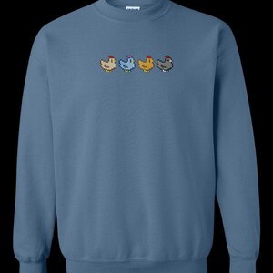 K&ouml;nnte beinhalten: Ein blaues Sweatshirt mit einer Reihe von f&uuml;nf gestickten, pixelartigen H&uuml;hnern in verschiedenen Farben.